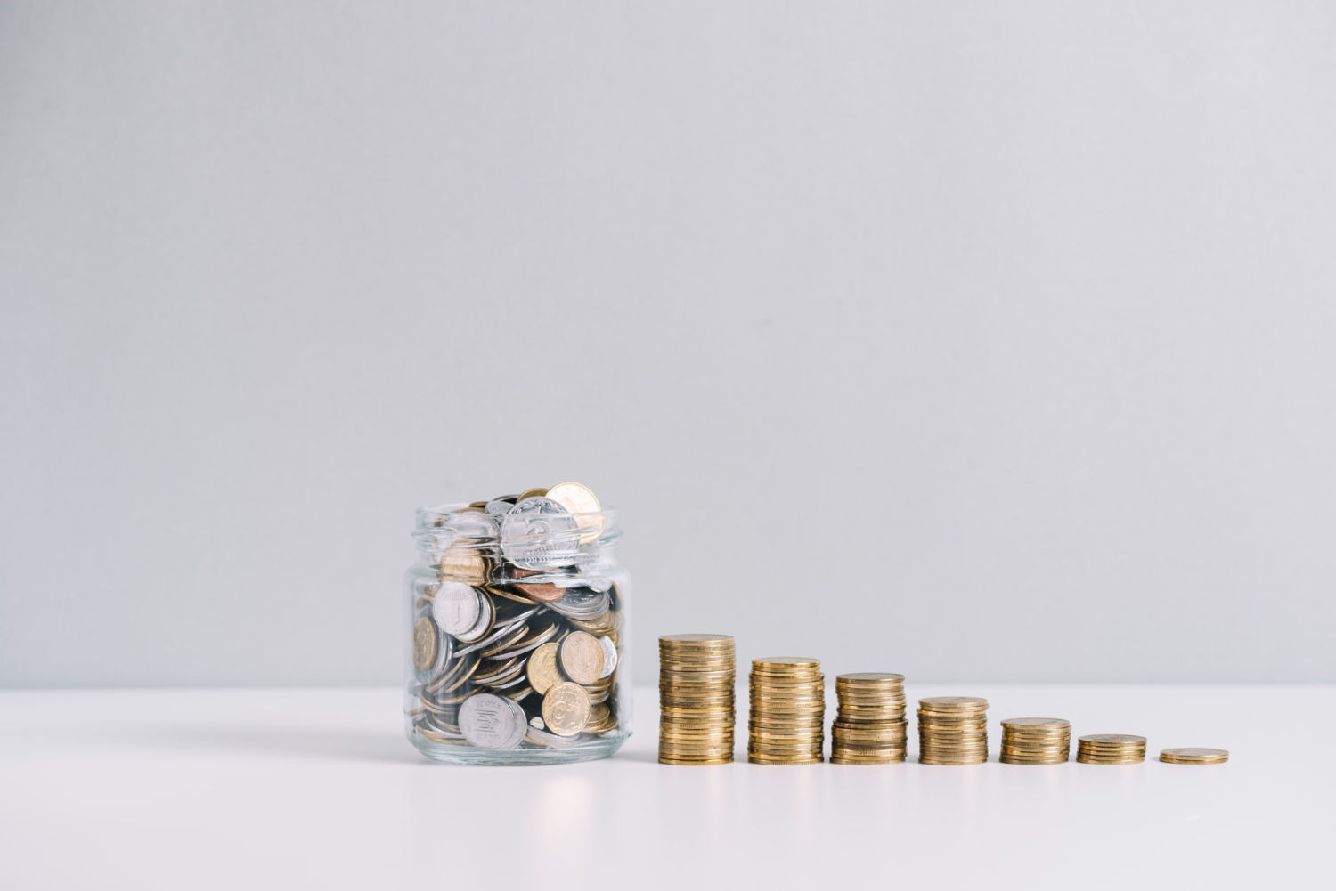 A glass jar filled with mixed coins beside stacked coin piles on a white surface.