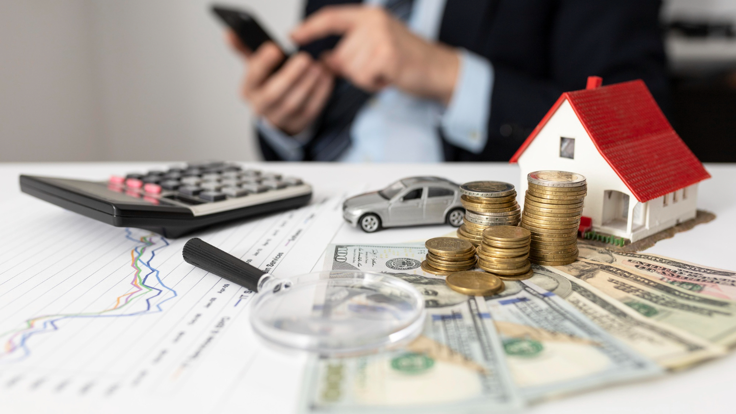 Businessman using mobile phone with financial documents, model car, house, and stacks of coins on desk.