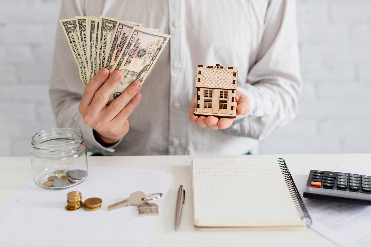 A person holds cash in one hand and a wooden house model in the other, with coins, a notebook.