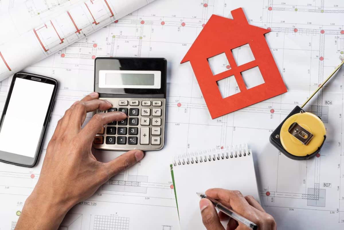 Hands calculating on a calculator beside architectural plans, a house model, and measuring tools on a desk.