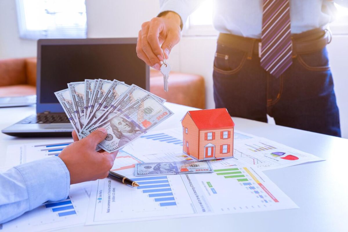 Two businessmen exchanging house keys and money over a desk with financial documents and a small house model.