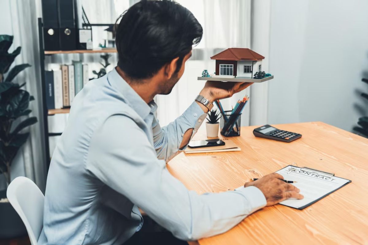A man in a dress shirt sits at a desk holding a model house, with a contract, calculator, and office supplies on the table.