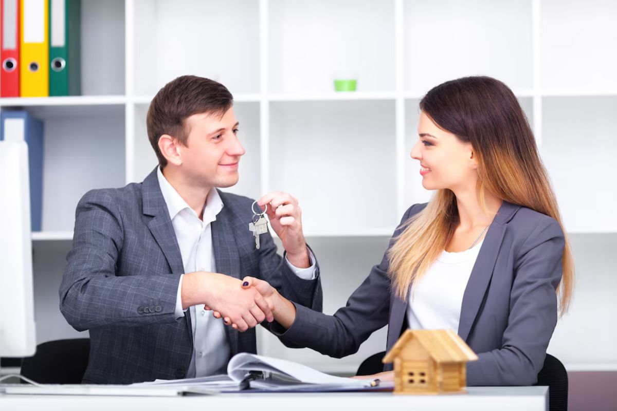 A man hands keys to a woman while they shake hands across a desk in an office, suggesting a successful real estate or business transaction.