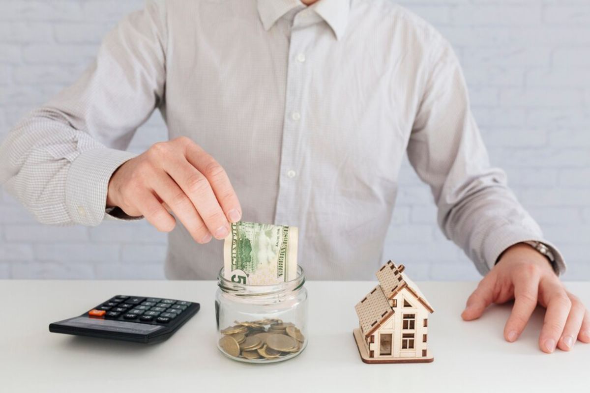 A person in a button-up shirt drops a $5 bill into a jar of coins beside a miniature wooden house and a calculator.