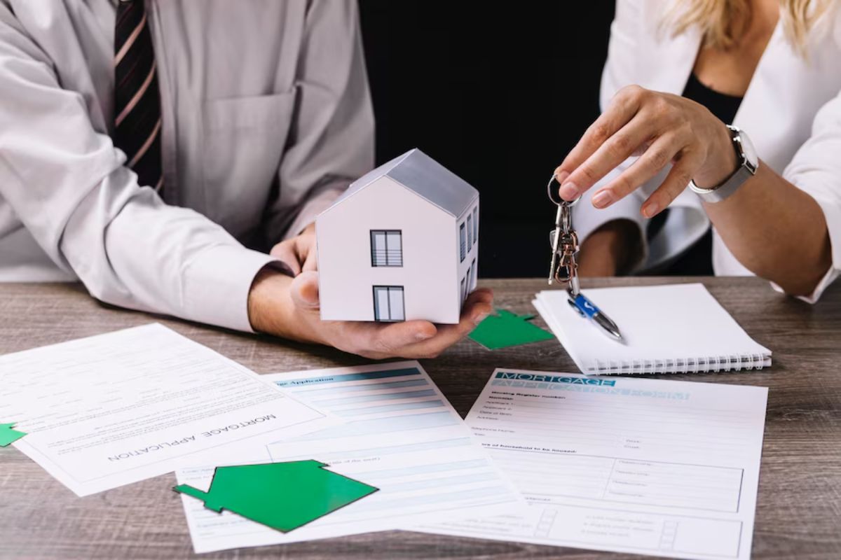 Two professionals at a desk with documents, a model house, and house keys, symbolizing a real estate transaction.
