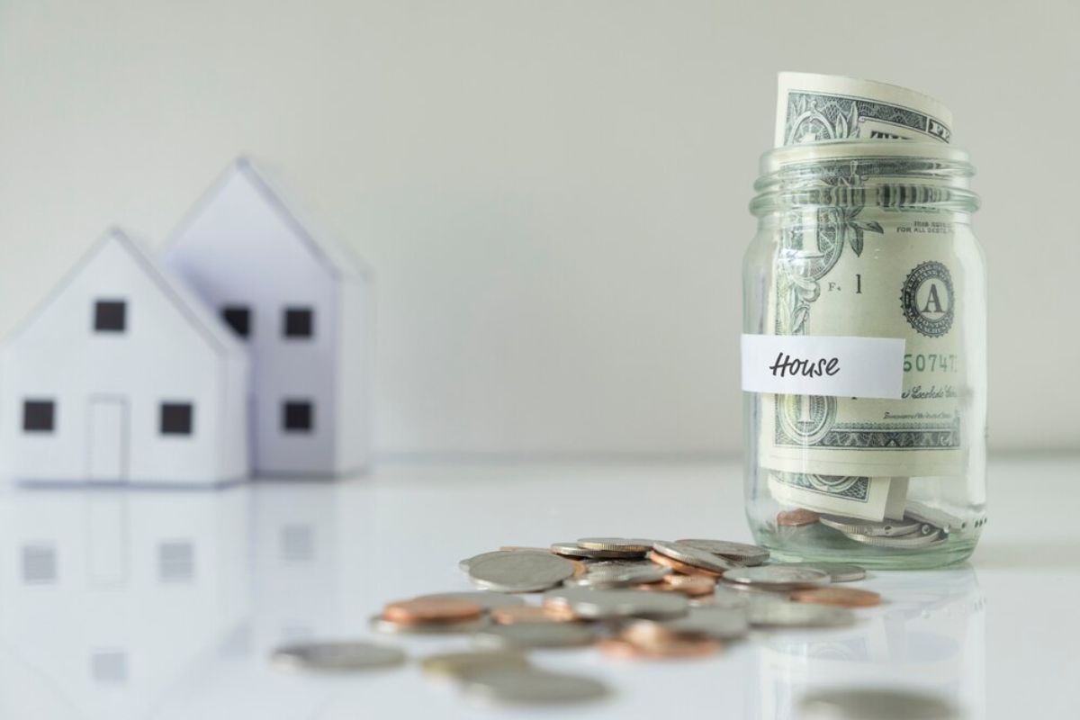 A glass jar labeled House filled with dollar bills, with coins scattered nearby and mini house models in the background.