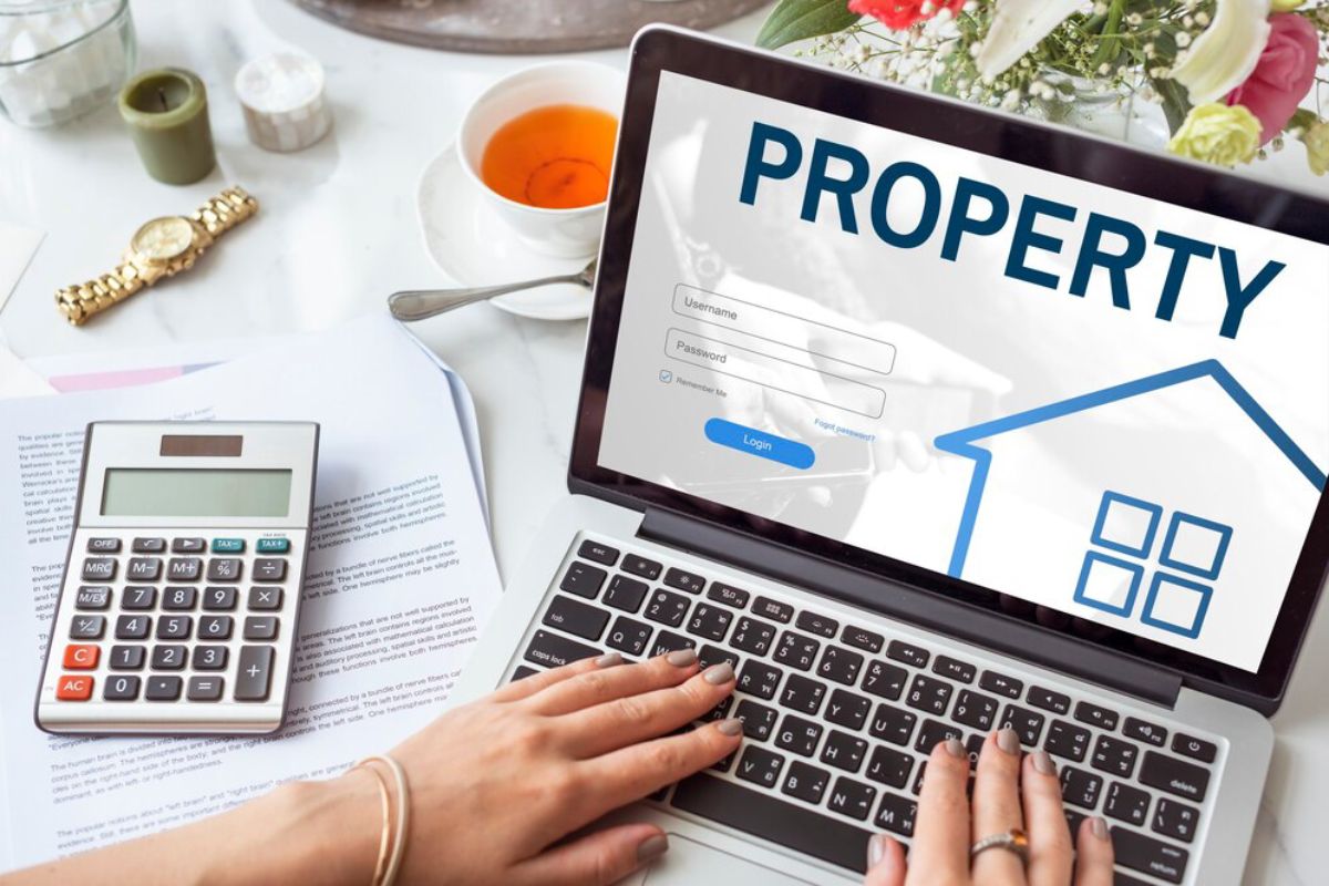 Woman using laptop with property website onscreen, calculator, paperwork, and a cup of tea on desk.