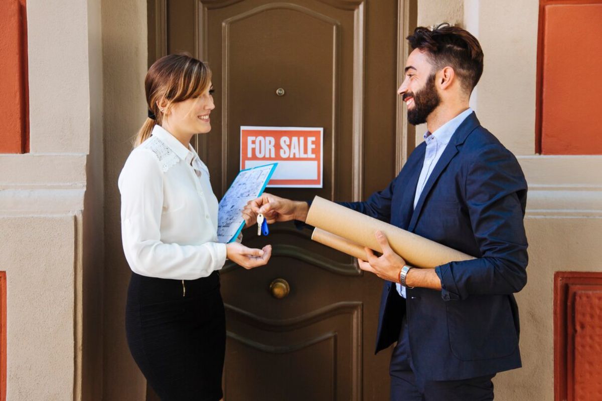 A man hands a woman house keys in front of a door with a For Sale sign.