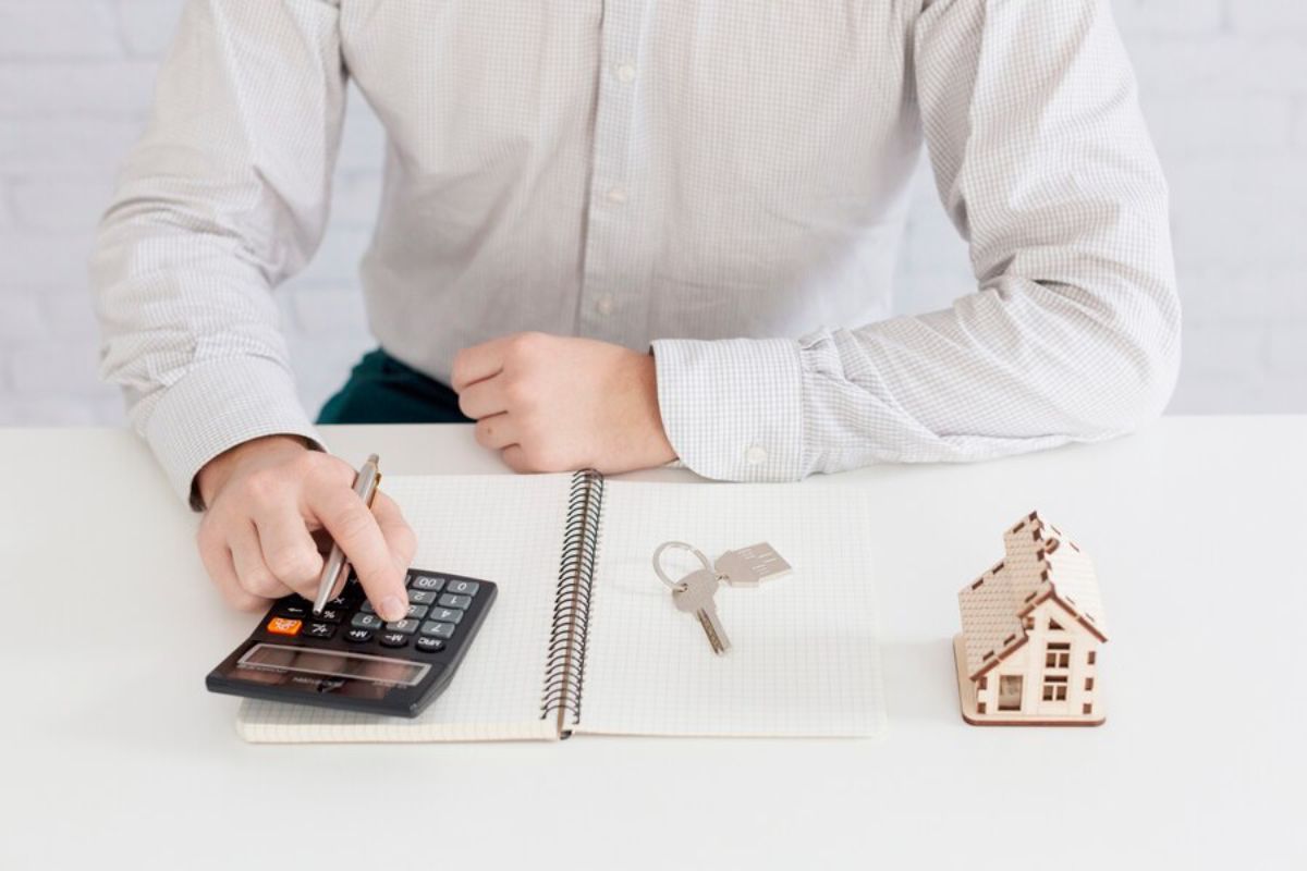 Man calculating finances with a calculator, notebook, keys, and miniature house on a desk.