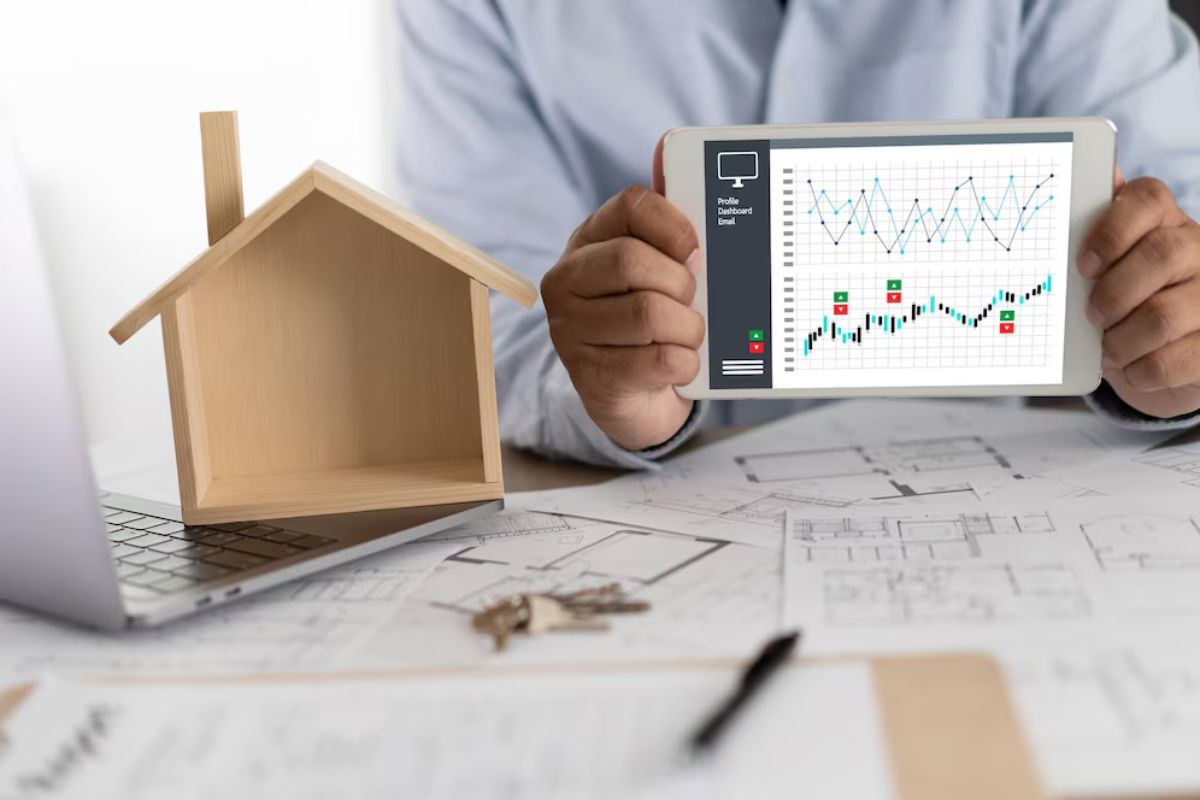 A person analysing financial graphs on a tablet with a wooden house model and blueprints on the table.
