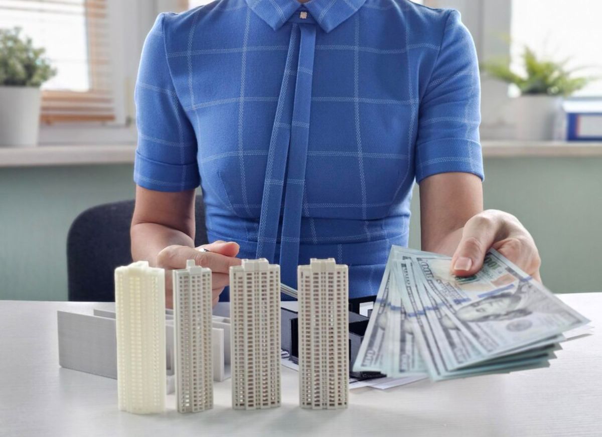 Person at desk arranging miniature buildings and handling US dollar bills, suggesting a financial or real estate business concept.