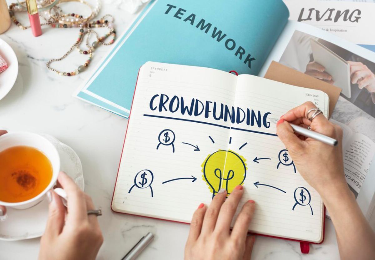 Person writing 'Crowdfunding' in a notebook, illustrating concept with bulb and money icons, surrounded by tea and accessories.