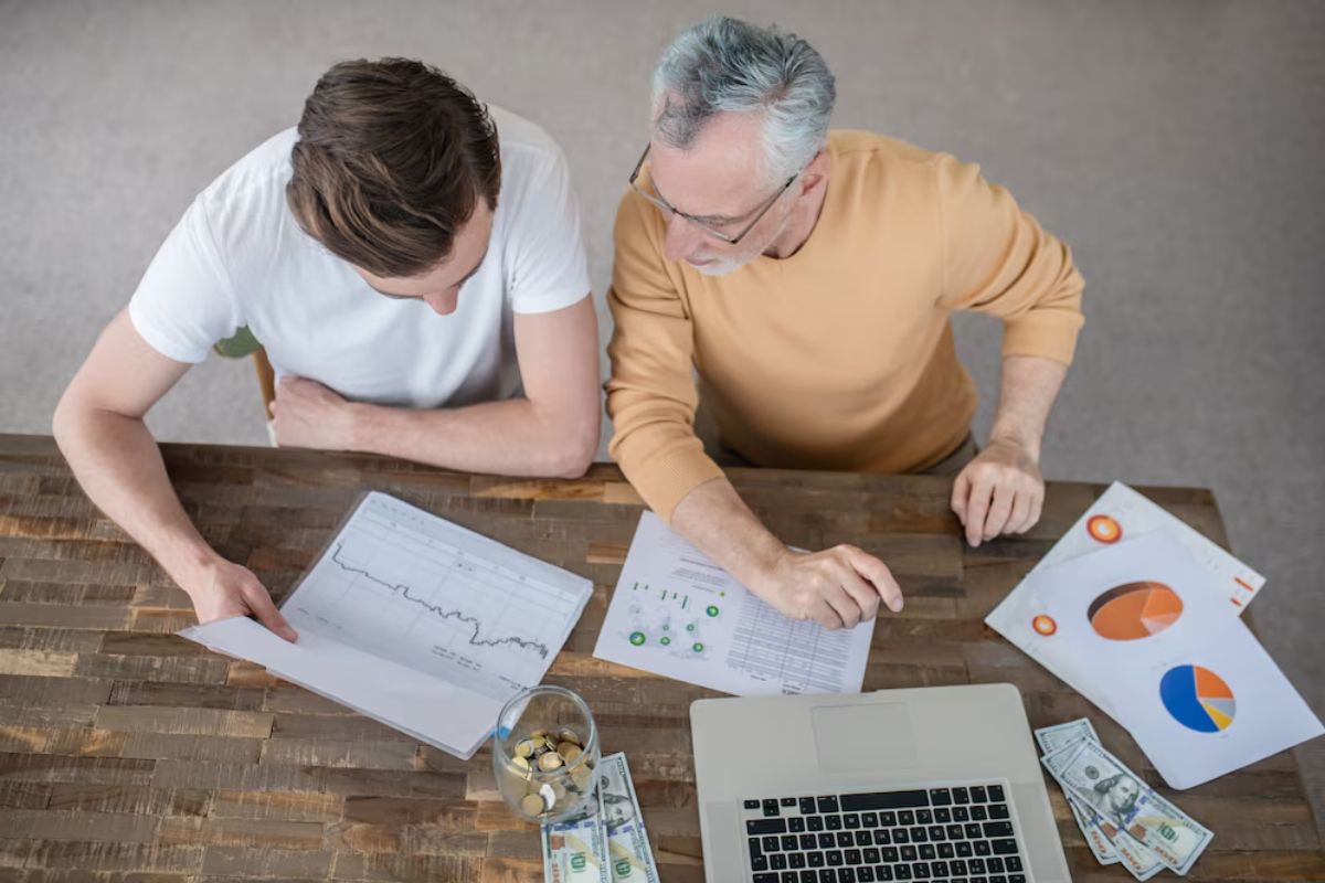 Two men examining financial documents and charts at a wooden table with a laptop and money.