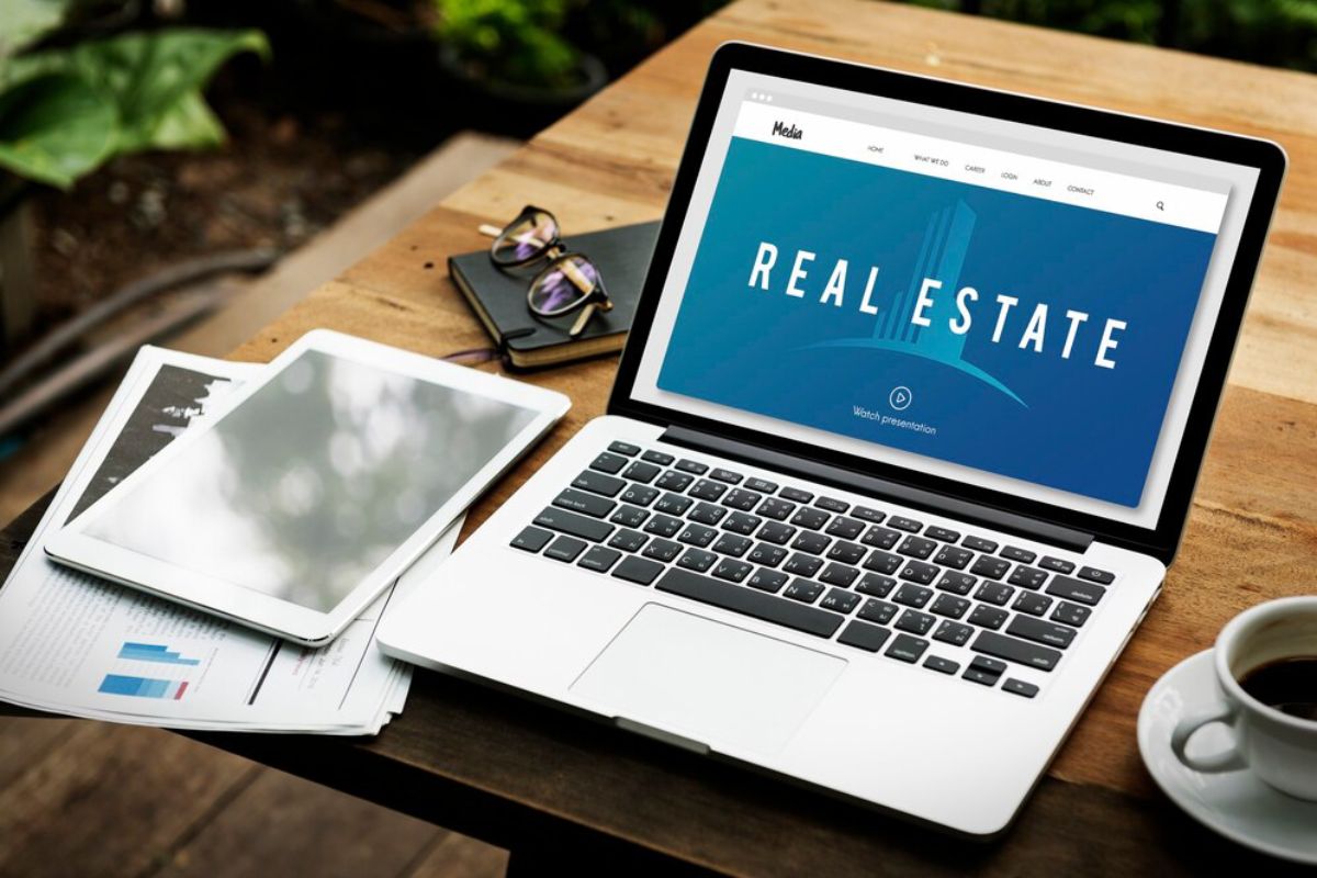 A laptop on a wooden table displays a real estate website, surrounded by documents, a tablet, sunglasses, and a cup of coffee.