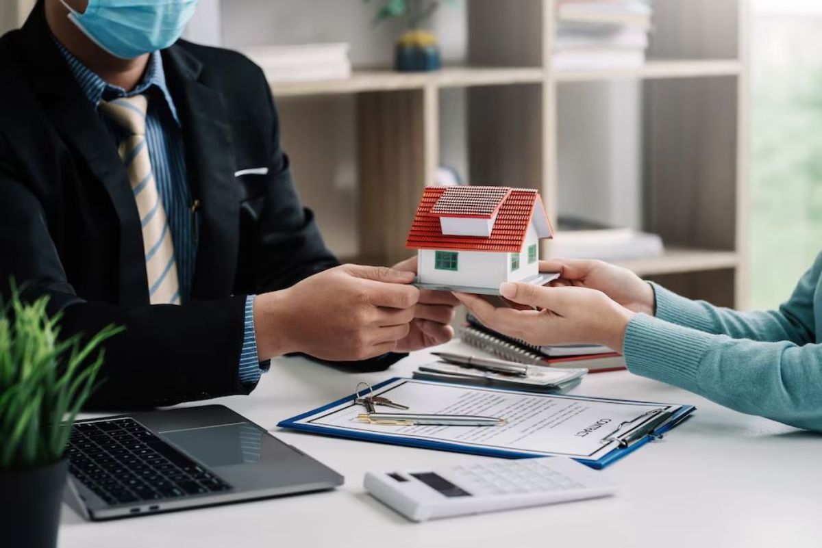 Two individuals at a desk, exchanging a model house, documents and laptop visible, both wearing masks.