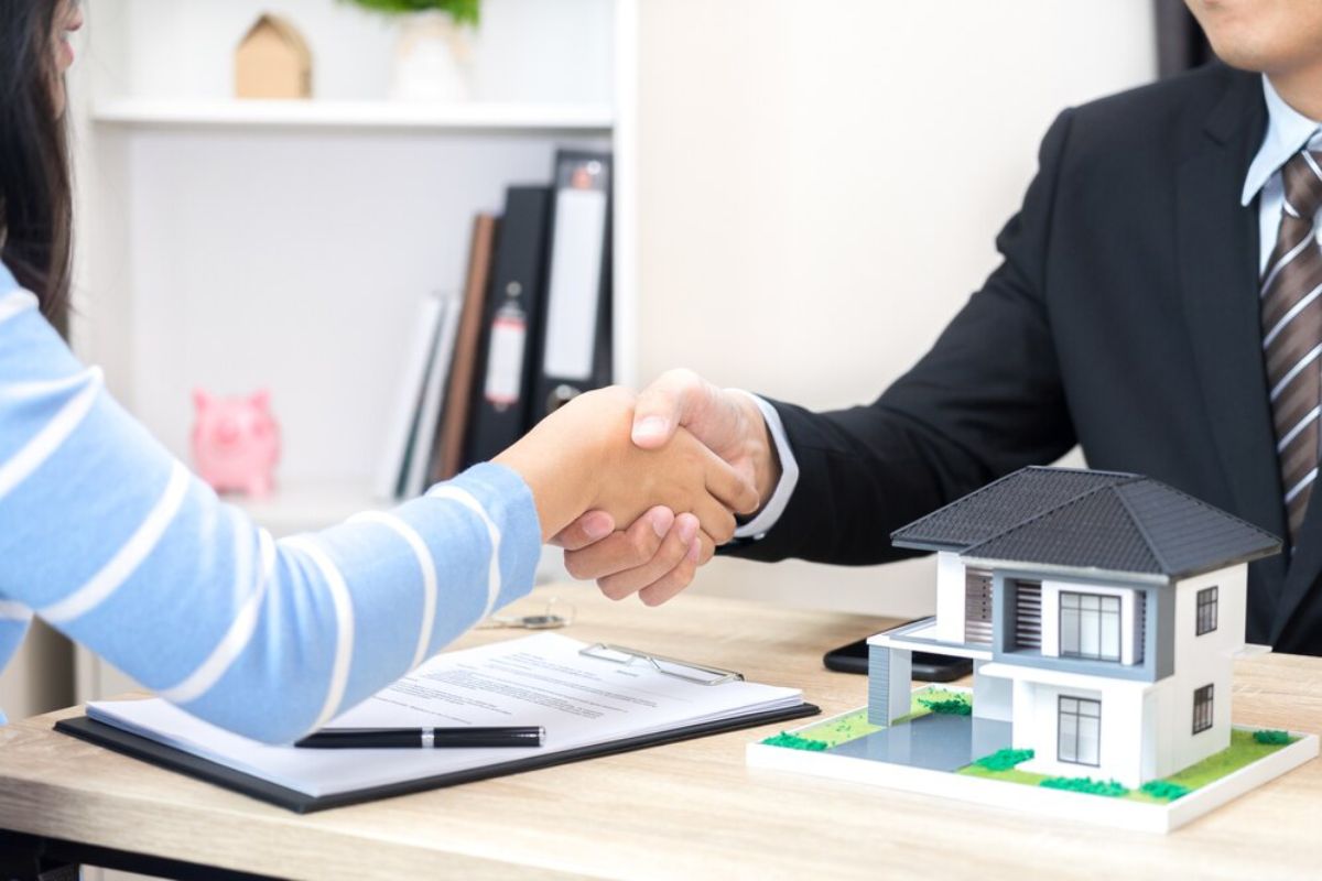 Two people shaking hands over a desk with a model house, symbolizing a real estate agreement.